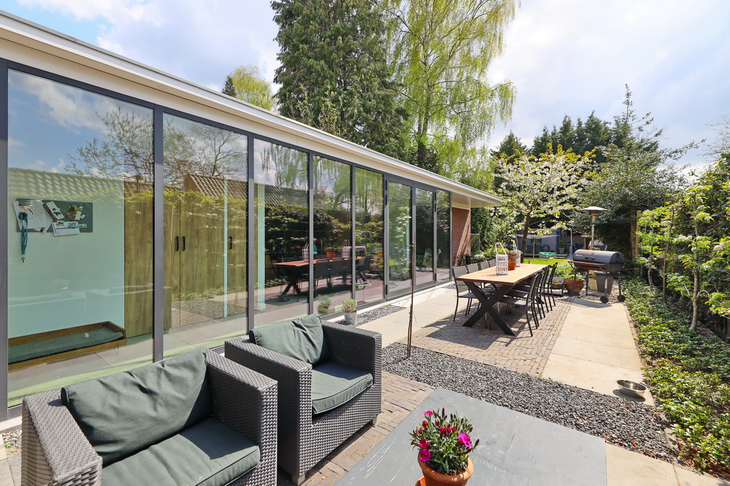 Diepe tuin met een moderne aanbouw. De glazen ramen van de aanbouw met de keuken, weerspiegelen de de tuin en de tuinmeubels.